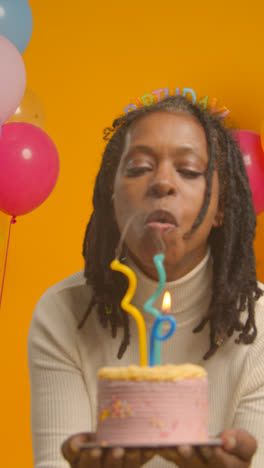 Vertical-Video-Studio-Portrait-Of-Woman-Wearing-Birthday-Headband-Celebrating-Birthday-Blowing-Out-Candles-On-Cake